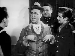 a man in what appears must be a brightly colored suit and hat, as this is a black and white photo, is the centre of attention speaking to three young men