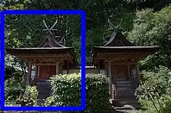 A small wooden building next to another identical building with gabled roof, a stair on the gable side covered by an extended roof. The roof ridge has forked finials.
