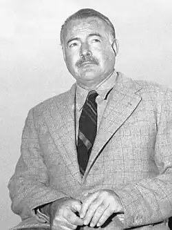 Dark-haired man in light colored short-sleeved shirt working on a typewriter at a table on which sits an open book