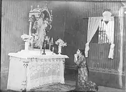 Black and white photo showing a young woman kneeling, praying before an altar with a statue of the Virgin Mary.