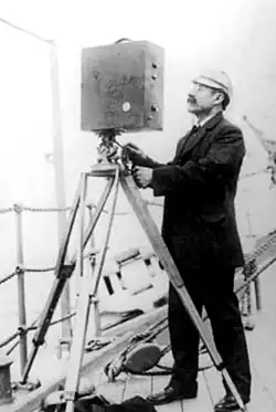 Eugenio Py, a pioneer of Argentine filmmaking, stands full-body in a black-and-white photo, next to a vintage camera