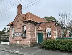 An old brick library with a brown roof and a large brick chimney. A sign on the side reads "THE OLD LIBRARY"