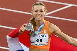 Photo of Femke Bol holding a silver medal in her hand and a Dutch flag behind her back with a red athletics track in the background