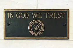 A gold-and-bronza plaque. It has a gold frame, and in the center of the plaque is the United States seal and the words "IN GOD WE TRUST" engraved in gold.