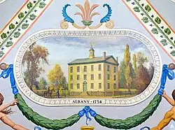 A painting of a yellow house surrounded by trees. Below the house is a garland with blue ribbons.