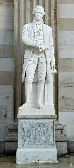 A white marble statue of Alexander Hamilton, who is in a coat and skinny pants. He is holding his right hand to his chest, and the statue is on a grey marble pedestal.
