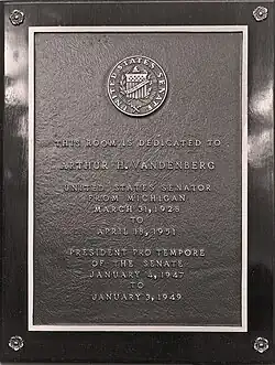 A black-and-gold plaque. On the plaque is text that reads "THIS ROOM IS DEDICATED TO ARTHUR H. WANDENBURG, UNITED STATES SENATOR FROM MICHIGAN", and text below that is unable to be distinguished from the plaque itself.