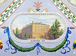 A painting of a building in Baltimore, Maryland. The building, yellow in color with several windows lining it's exterior walls, is surrounded by trees. Below the painting is a painted garland with blue ribbons, and directly below the building is the text "BALTIMORE, 1776".