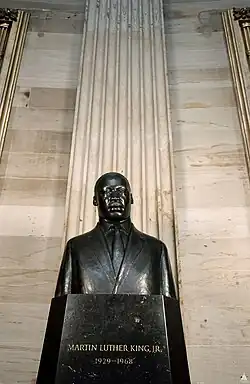 A black bronze statue of Dr. Martin Luther King Jr. He is wearing a suit, and is standing behind a lectern, presumably depicting him during a speech. Rising behind him is a large pillar.