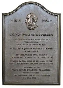 A bronze plaque. On the center-top is a sideview of someone's head, and the largest part of the plaque reads "CANNON HOUSE OFFICE BUILDING PURSUANT TO PUBLIC LAW 37-453 APPROVED MAY 21, 1963 THIS BUILDING WAS NAMED IN HONOR OF THE HONORABLE JOSEPH GURNEY CANNON". Smaller parts of the plaque are unintelligible, due to lighting.