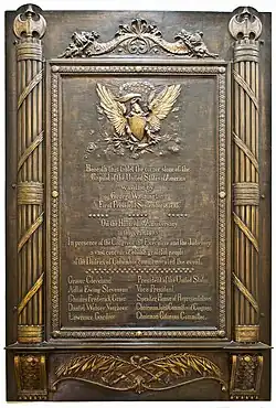 A bronze plaque with engraved pillars on the right and left. Atop the pillars are eagles, and in the middle of the plaque is another eagle engraving. The plaque reads "Beneath this tablet the corner stone of the Capitol of the United States of America was laid by George Washington First President September 18, 1793".