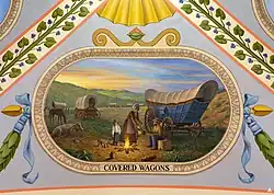 Two different types of covered wagons are shown, along with livestock and a family of settlers cooking a meal over an open fire.