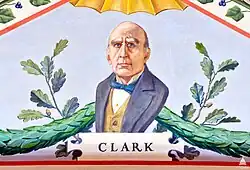 A painting depicting a portrait of David Lynn. He is wearing a grey suit with a blue bowtie, and is looking directly forward. Behind him are green garlands and plants, and directly under him is the text "CLARK".