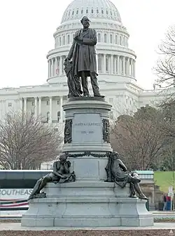 The tapered, cylindrical granite pedestal holds four over-life-size bronze figures, with the portrait statue of Garfield at the top and three allegorical figures representing different phases of his career below. Garfield is depicted in giving a speech, gazing intently outward with a sheaf of papers in his left hand, his right hand rests on a book on a draped column. The toe of one shoe projects over the edge of the base, giving the work a sense of vigor and movement.