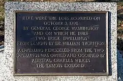 A black-and-gold bronze plaque held up by a white granite block. On the plaque, text reads: "HERE WERE THE LOTS ACQUIRED ON OCTOBER 3, 1798 BY GENERAL GEORGE WASHINGTON AND ON WHICH HE BUILT TWO BRICK DWELLINGS FROM DESIGNS BY DR. WILLIAM THORTON A DWELLLING REMODELED FROM THE TWO DWELLINGS WAS OWNED AND OCCUPIED BY ADMIRAL CHARLES WILKES THE FAMOUS EXPLORER".