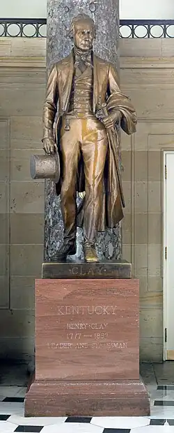 A brown bronze statue of Henry Clay. In his right hand is a top hat, and in his left hand is a cane. On the upper part of the pedestal is the word "CLAY" engraved, and on the main pedestal are the words "KENTUCKY, HENRY CLAY".