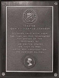 A brown-and-grey bronze plaque. On the bottom of the plaque is a sideview of John F. Kennedy, and the plaque reads: "SENATOR JOHN FITZGERALD KENNEDY OCCUPIED THIS ROOM FROM THE TIME HE WAS NOMINATED FOR OFFICE OF THE PRESIDENT OF THE UNITED STATES ON JULY 13, 1960 UNTIL HIS INAUGURATION ON JANUARY 20, 1961".