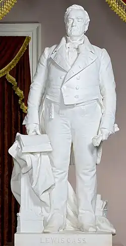 A white marble statue of Lewis Cass. He is holding a paper in is left and, and is wearing a suit. Engraved on the pedestal are the words "LEWIS CASS".