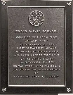 A grey-and-gold bronze plaque with the United States seal at top. The plaque reads: "LYNDON BAINES JOHNSON OCCUPIED THIS ROOM FROM JANUARY 7, 1959, TO NOVEMBER 22, 1963; FIRST AS MAJORITY LEADER OF THE UNITED STATES SENATE AND LATER AS VICE PRESIDENT OF THE UNITED STATES. ON NOVEMBER 22, 1963, HE WAS SWORN IN AS PRESIDENT FOLLOWING THE ASSASSINATION OF PRESIDENT JOHN F. KENNEDY".