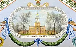 A yellow brick building surrounded by trees. Below the building are green garlands with blue ribbons, and directly under the building are the words "PHILADELPHIA, 1775".