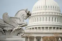 A white marble winged horse, looking down and shown in the middle of a prance. On the right is the dome of the Capitol, out of focus.