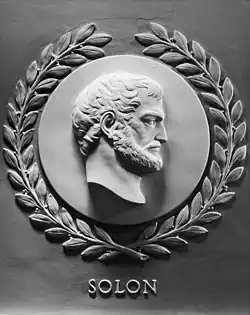 A grey marble sideview of Solon. He is looking to the right, and has a beard. Surrounding the circular frame is a laurel wreath. Under the engraving is the text "SOLON".