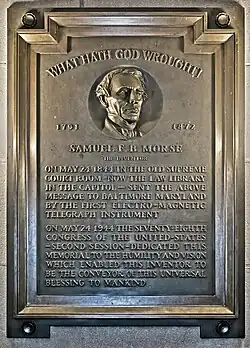 A brown bronze plaque with the face of Samuel L. Morse engraved into it. The text on the plaque reads: "WHAT HATH GOD WROUGHT! 1791 1872 SAMUEL F.B. MORSE THE INVENTOR ON MAY 24 1844 IN THE OLD SUPREME COURT ROOM - NOW THE LAW LIBRARY IN THE CAPITOL - SENT THE ABOVE MESSAGE TO BALTIMORE MARYLAND BY THE FIRST ELECTRO-MAGNETIC TELEGRAPH INSTRUMENT ON MAY 24 1944 THE SEVENTY-EIGHTH CONGRESS OF THE UNITED-STATES -SECOND SESSION - DEDICATED THIS MEMORIAL TO THE HUMILITY AND VISION WHICH ENABLED THIS INVENTOR TO BE THE CONVEYOR OF THIS UNIVERSAL BLESSING TO MANKIND".
