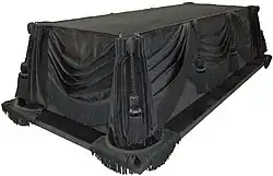 A rectangular wooden coffin draped with a black cloth.