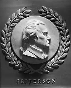 A grey marble sideview of Thomas Jefferson. He is looking to the right, and is smiling. Surrounding the circular frame is a laurel wreath. Under the engraving is the text "JEFFERSON".