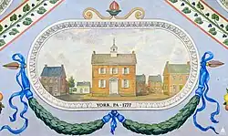 After leaving Baltimore the Congress met briefly in Philadelphia but soon moved to York, Pennsylvania, where it met for nine months in the old Court House. The building in the center of the image is the Court House, and under it is a green garland with blue ribbons. Surrounding the court are several smaller buildings.