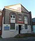 Close-up front view of a cobbled flint building with brick dressings and a white-painted area around the entrance door, which is reached up a ramp. The lower storey has two sash windows; these are linked to the upper storey's two semicircular-headed windows by areas of white panelling. The building is topped by a flint pediment with a semicircular louvre.