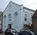 Three-quarter view of a pale blue building with a plain exterior and a large pediment, on a crowded site between two flint structures. The façade has three tall, triple-recessed blank arches, the centre of which is shortened to accommodate an entrance door. A round opening sits just below the pediment. The side has round-arched windows.