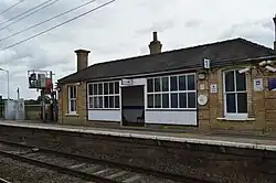 A small single-story yellow brick building. The photograph shows the platform side from the other building. One of the tracks below the platform, the overhead wires, and a signal are also visible in the photo.