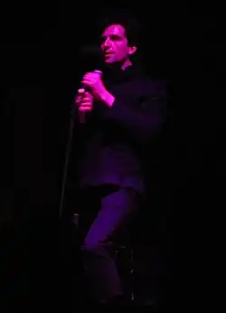 Francis Farewell Starlite partially illuminate on a darkened stage, holding a microphone