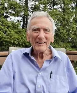 Elderly caucasian male with grey hair in shirtsleeves sitting on the terrace which looks onto woods