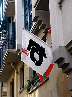 A short mast with a plastic flag hanging from a building. The flag has a white background, a black symbol with bold strokes and a hollow inside. The flag also has the letters "nistia" partially visible in red.