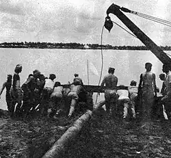 Black and white photograph of a group of people pushing a structure into a river with the assistance of a crane