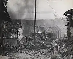 two American soldiers running from left to right through a burning village