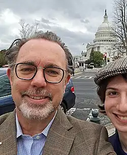 close-up of Garett Jones wearing a light blue checkered shirt and a tan checkered jacket, grinning directly at camera, which he appears to be holding, with younger person to the left, partly out of frame; US Capitol is in background