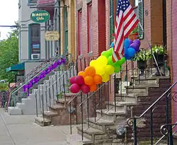 Stoops on some brick houses, seen looking along the sidewalk. One has balloons in the colors of the rainbow tied to its railing. Another has a purple ribbon.