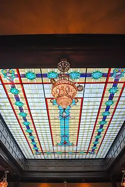 View looking up at the skylight. I'm guessing it's about 10 feet by 20. There is a chandelier in the foreground. The skylight is rectangular. The outer row of panes are white with gold stripes, the next row in are gold squares with turquoise rosettes and a navy blue band running through each forming a rectangle. The next band in is a repeat of the outer white band. The inner section is a rectangle with a long turquoise column-shaped center piece.