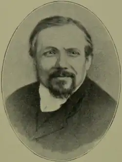 A vintage oval portrait of a man with short hair, a moustache, and a goatee, wearing a high-collared shirt and a dark coat, smiling slightly at the camera.