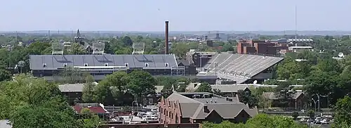 An elevated view of several buildings and the trees surrounding them. A red brick building with a sloped roof is in the foreground, and a large white football stadium is just behind it, taking up much of the center of the picture. Beyond the stadium, there is a red brick smokestack near the center of the picture, the red brick Tech Tower building on the left side bearing white letters that spell "TECH", and the red brick physics building on the right side. In the background there is a white domed building. All around these buildings are green-leafed oak trees. An overcast, light blue sky takes up the top third of the picture.