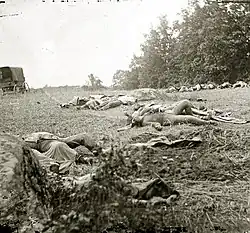 Square black-and-white photograph showing bodies on the ground. A wagon can be seen on the left.