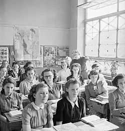 Black-and-white photo of a highschool classroom of girls looking to the front while their teacher stands behind them and explains