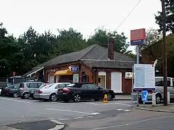 A single-story red brick building as viewed from across the street. The view of the building is blocked by many cars parked in front and a sign at the entrance to the car park. Behind the sign is a pole, at the top of which there is a double arrow sign with "Gordon Hill" written beneath it.