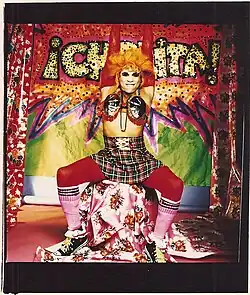 Flashy drag queen sits on a floral-draped stump on a stage with her arms raised in the air, in front of a colorfully decorated DIY background
