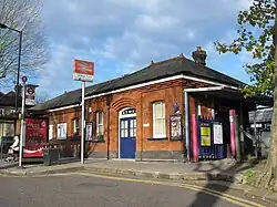 A single storey red-brick building as photographed from the road. It has a large blue door which is closed, and instead the entrance is through a gate to the right. In front of the building is a bus stop and a double arrow on a pole with a sign underneath that reads "Grange Park".