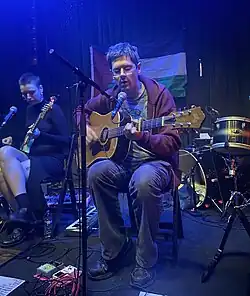 A man and a woman performing on acoustic guitar and bass respectively in front of a Palestinian flag