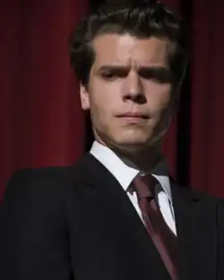 Photograph of Gus Halper as Erik Menendez in a suit, staring pensively at what can be presumed to be the ground.